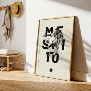May include: Framed black and white poster featuring a soccer player in action, with the name "MESSI" and the number "10" in large, bold text. The poster has a light beige background and a wooden frame.
