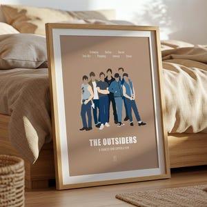 May include: A framed poster featuring a group of stylized figures from "The Outsiders" film. The poster has a tan background with the film title and character names at the top. The frame is a light wood color.