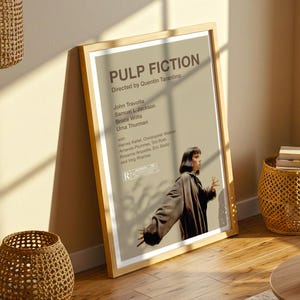K&ouml;nnte beinhalten: Ein gerahmtes Filmplakat f&uuml;r "Pulp Fiction" unter der Regie von Quentin Tarantino. Das Plakat zeigt den Filmtitel, die Besetzung und den Abspann in einem beige-wei&szlig;en Farbschema. Das Bild zeigt eine Frau in einem braunen Mantel.