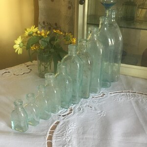 Puede incluir: Una colecci&oacute;n de ocho botellas de vidrio transparente con un ligero tono aguamarina, dispuestas en l&iacute;nea diagonal por tama&ntilde;o, de menor a mayor. Cada botella presenta un cuello corto y un cuerpo redondeado, con variaciones sutiles en su forma.