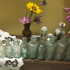 May include: A collection of vintage glass bottles with a variety of colors and shapes, arranged on a wooden shelf. The bottles are displayed on a white lace tablecloth with a floral arrangement in a brown glass bottle.