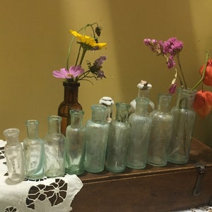 May include: A collection of vintage glass bottles with a variety of dried flowers in them. The bottles are arranged on a wooden shelf with a white lace tablecloth in front.