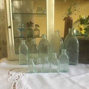 Puede incluir: Una colecci&oacute;n de diez botellas de vidrio transparente y de tono azul claro de diversos tama&ntilde;os y formas, dispuestas en dos filas. Las botellas presentan cuellos estrechos y cuerpos redondeados, mostrando algunas texturas superficiales sutiles.
