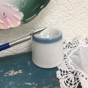 Puede incluir: Un plato de cerámica blanco con un borde azul, un pincel plateado y azul y un encaje blanco sobre una superficie de madera azul.