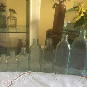 Puede incluir: Una colecci&oacute;n de nueve botellas de vidrio transparente con un ligero tono azulado, dispuestas en fila por tama&ntilde;o, de menor a mayor. Cada botella presenta un cuello estrecho y un cuerpo redondeado, mostrando una est&eacute;tica vintage con texturas.