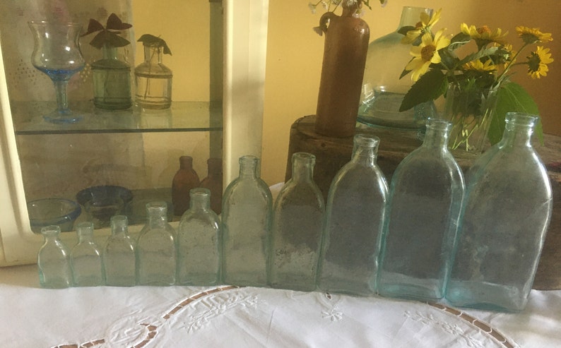 Puede incluir: Una colecci&oacute;n de nueve botellas de vidrio transparente con un ligero tono azulado, dispuestas en fila por tama&ntilde;o, de menor a mayor. Cada botella presenta un cuello estrecho y un cuerpo redondeado, mostrando una est&eacute;tica vintage con texturas.