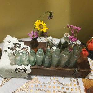 May include: A collection of small, clear glass bottles with cork stoppers, arranged on a wooden box. The bottles are filled with dried flowers, including yellow sunflowers, pink cosmos, and purple flowers. The box is sitting on a white lace tablecloth.