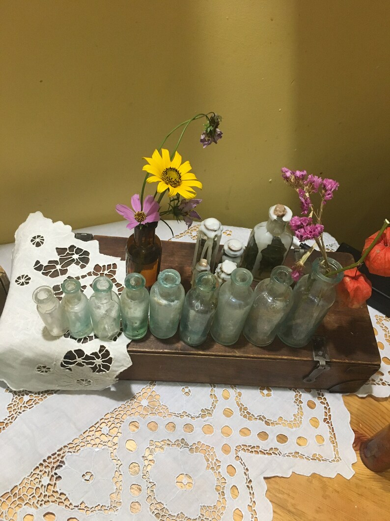 May include: A collection of small, clear glass bottles with cork stoppers, arranged on a wooden box. The bottles are filled with dried flowers, including yellow sunflowers, pink cosmos, and purple flowers. The box is sitting on a white lace tablecloth.