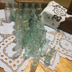 May include: A collection of vintage glass bottles with a light blue tint. The bottles are arranged in a pyramid shape on a white lace tablecloth.