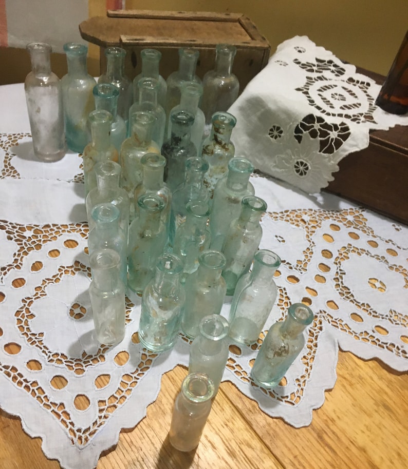 May include: A collection of vintage glass bottles with a light blue tint. The bottles are arranged in a pyramid shape on a white lace tablecloth.