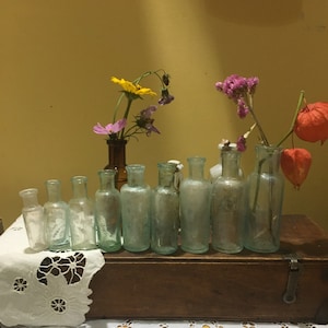 May include: A collection of ten clear glass bottles with a variety of dried flowers in them. The bottles are arranged in a row on a wooden box with a white lace tablecloth in front of it.