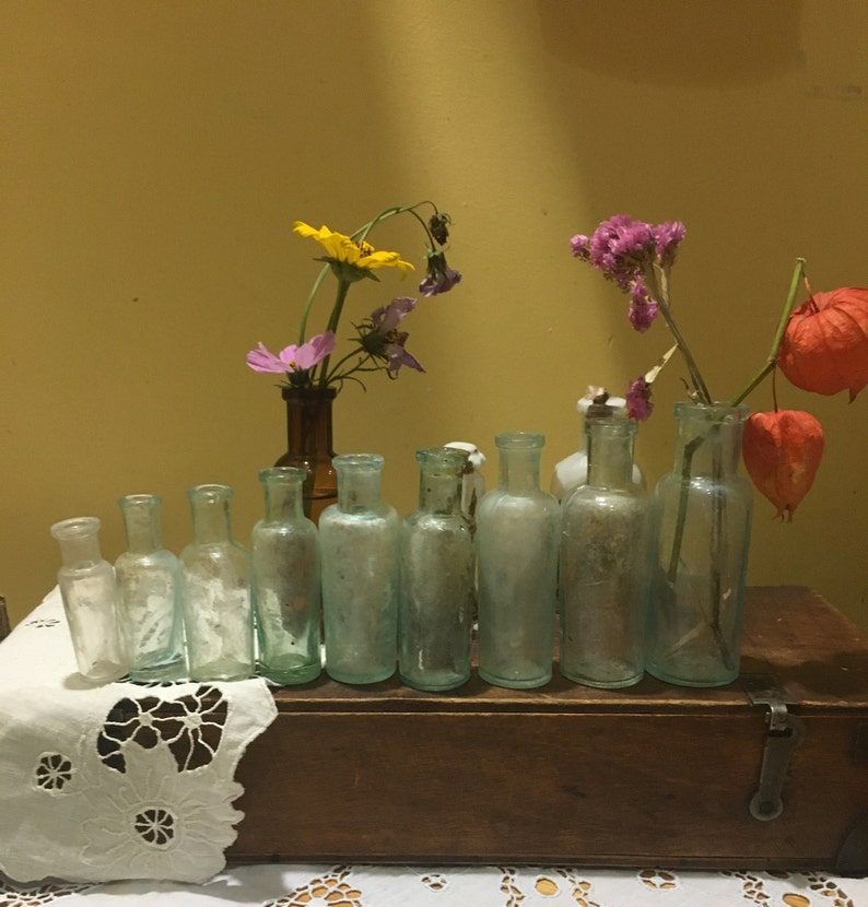 May include: A collection of ten clear glass bottles with a variety of dried flowers in them. The bottles are arranged in a row on a wooden box with a white lace tablecloth in front of it.