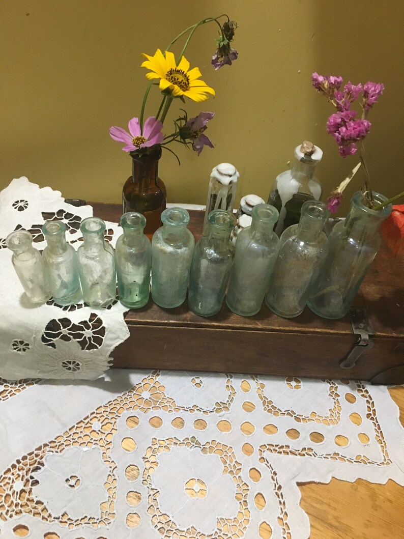 May include: A collection of vintage glass bottles with a variety of colors and shapes, arranged on a wooden shelf. The bottles are displayed on a white lace tablecloth with a floral arrangement in a brown glass bottle.