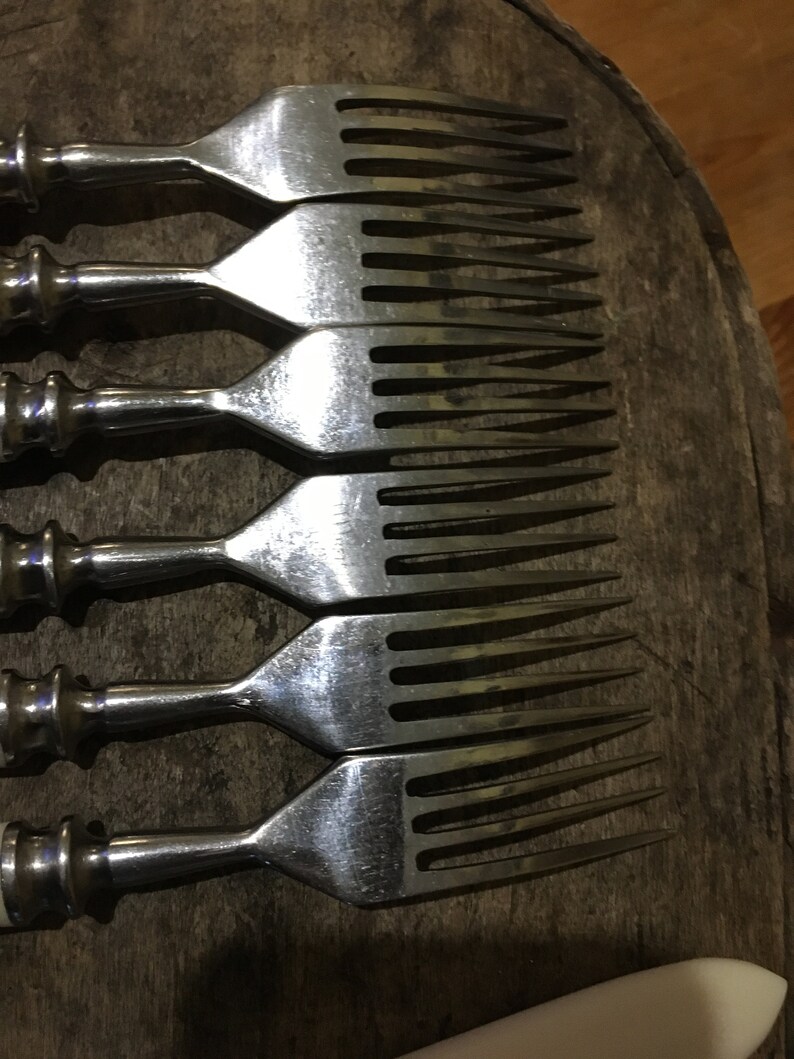 May include: Set of six silver forks with white handles. The forks are arranged in a row, with the tines facing up. The forks are on a wooden surface.
