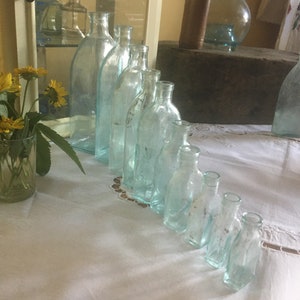 Puede incluir: Una colecci&oacute;n de nueve botellas de vidrio transparente con un ligero tono azulado, dispuestas en l&iacute;nea diagonal de mayor a menor. Cada botella presenta un cuello estrecho y una superficie lisa y transparente que refleja la luz.