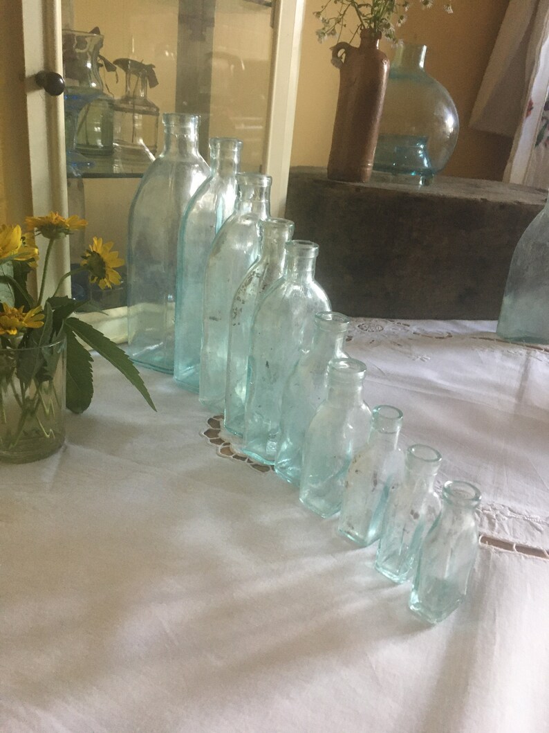 Puede incluir: Una colecci&oacute;n de nueve botellas de vidrio transparente con un ligero tono azulado, dispuestas en l&iacute;nea diagonal de mayor a menor. Cada botella presenta un cuello estrecho y una superficie lisa y transparente que refleja la luz.
