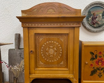 May include: A wooden cabinet with intricate carved floral and geometric designs. The cabinet has a small door with a keyhole and a decorative top. The wood has a warm, natural finish. A small shelf is below the door.