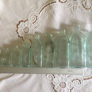 Puede incluir: Una colecci&oacute;n de nueve botellas de vidrio transparente de distintos tama&ntilde;os dispuestas en fila, de menor a mayor. Una regla de metal plateado est&aacute; colocada frente a las botellas, mostrando una longitud total de 30 cent&iacute;metros.