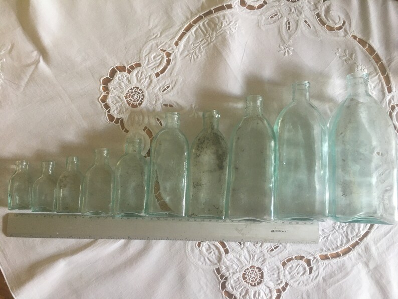 Puede incluir: Una colecci&oacute;n de nueve botellas de vidrio transparente de distintos tama&ntilde;os dispuestas en fila, de menor a mayor. Una regla de metal plateado est&aacute; colocada frente a las botellas, mostrando una longitud total de 30 cent&iacute;metros.