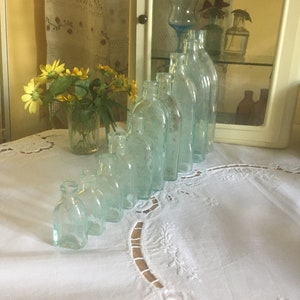 Puede incluir: Una colecci&oacute;n de nueve botellas de vidrio transparente con un ligero tono aguamarina, dispuestas en una l&iacute;nea diagonal por tama&ntilde;o, de menor a mayor. Cada botella presenta un cuello corto y un cuerpo redondeado con un aspecto antiguo.