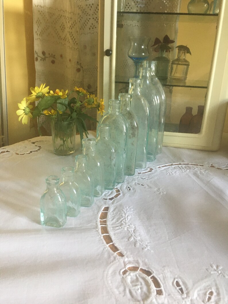 Puede incluir: Una colecci&oacute;n de nueve botellas de vidrio transparente con un ligero tono aguamarina, dispuestas en una l&iacute;nea diagonal por tama&ntilde;o, de menor a mayor. Cada botella presenta un cuello corto y un cuerpo redondeado con un aspecto antiguo.