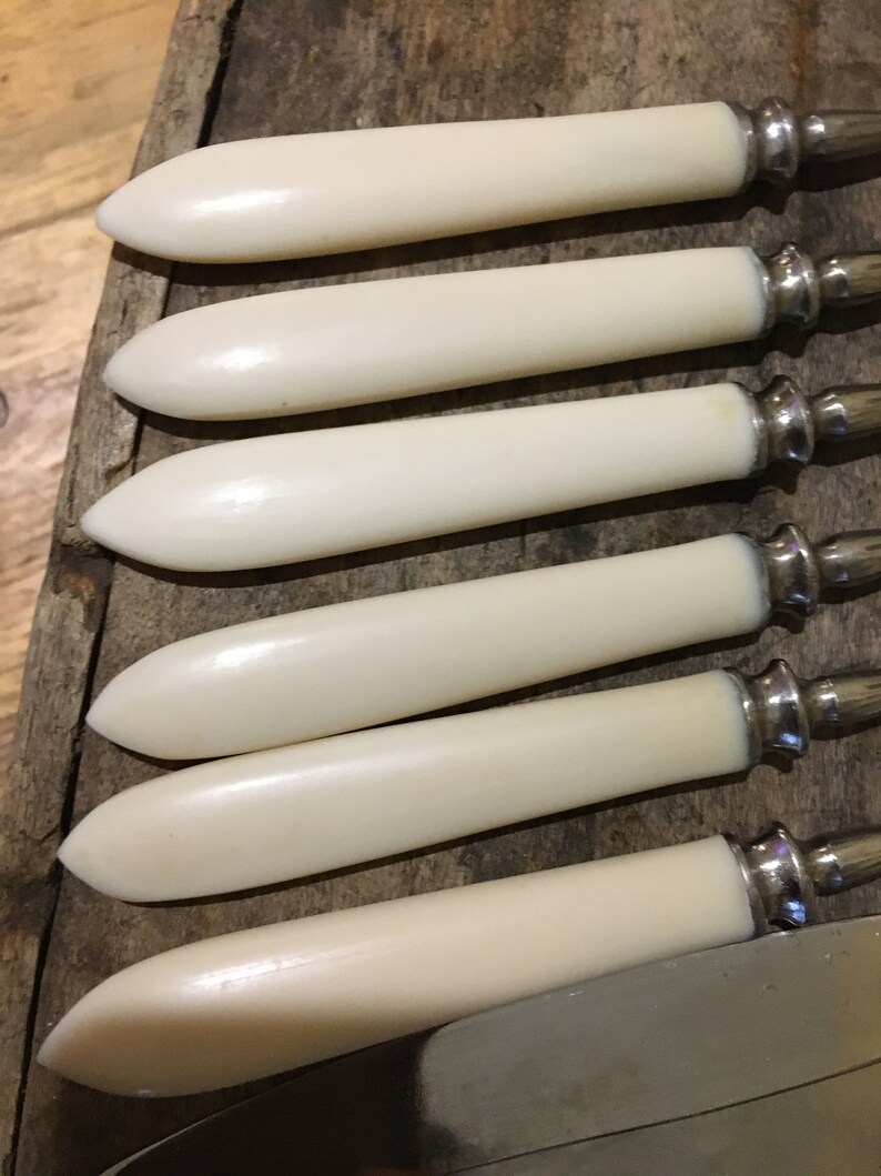 May include: Set of six ivory-colored butter knives with silver handles. The knives are arranged in a row on a wooden surface.