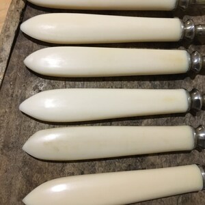 May include: Set of six ivory-colored butter knives with silver handles. The knives are arranged in a row, with the handles pointing to the right.