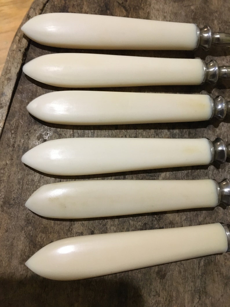 May include: Set of six ivory-colored butter knives with silver handles. The knives are arranged in a row, with the handles pointing to the right.