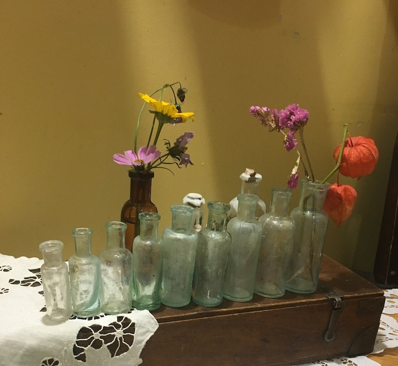 May include: A collection of vintage glass bottles with a variety of dried flowers in them. The bottles are arranged on a wooden shelf with a white lace tablecloth in front.