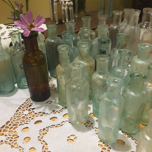May include: A collection of vintage glass bottles with a variety of shapes and sizes. The bottles are arranged on a white lace tablecloth. Some of the bottles are filled with a clear liquid, while others are empty.
