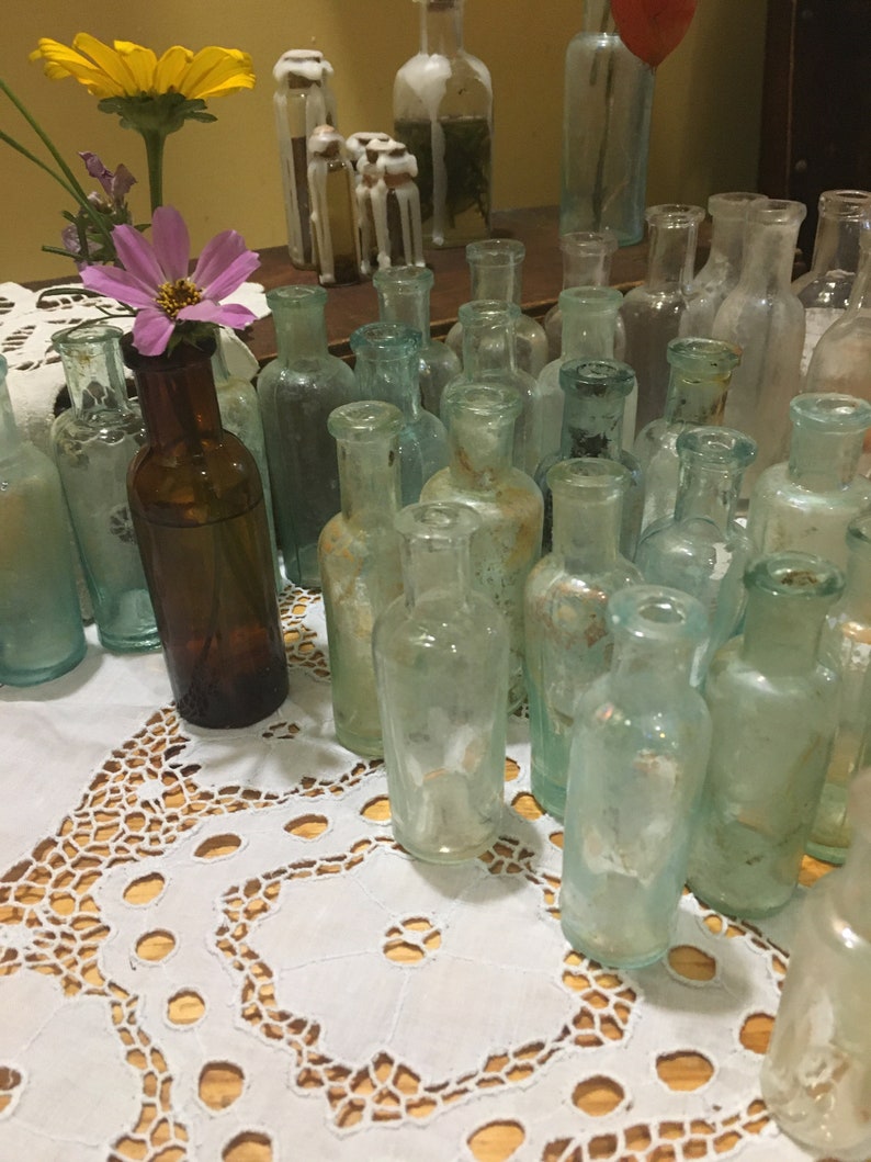May include: A collection of vintage glass bottles with a variety of shapes and sizes. The bottles are arranged on a white lace tablecloth. Some of the bottles are filled with a clear liquid, while others are empty.