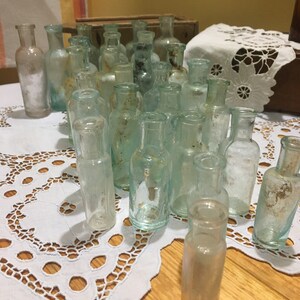 May include: A collection of vintage glass bottles in various shades of green and clear. The bottles are arranged in a triangular shape on a white lace tablecloth. The bottles are all different sizes and shapes, and some have a frosted or textured finish.