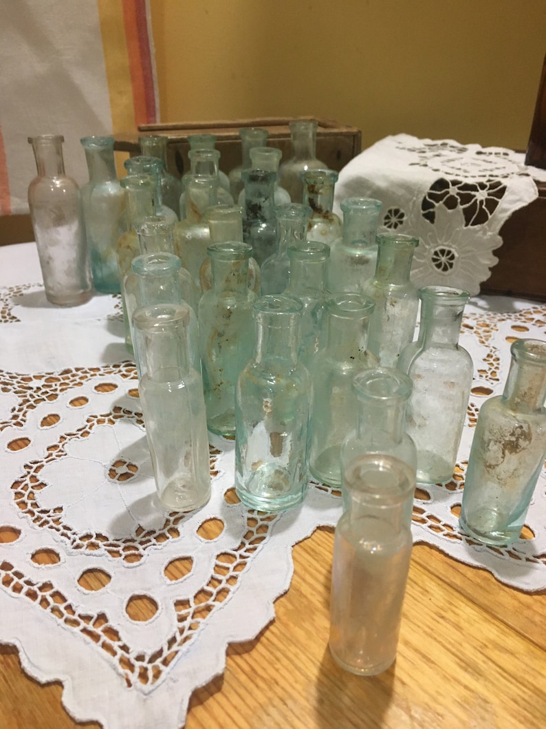 May include: A collection of vintage glass bottles in various shades of green and clear. The bottles are arranged in a triangular shape on a white lace tablecloth. The bottles are all different sizes and shapes, and some have a frosted or textured finish.