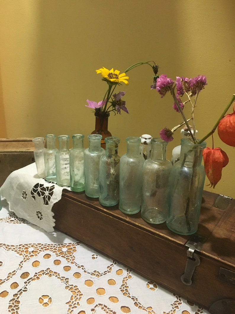 May include: A wooden box filled with clear glass bottles, each containing a single flower. The flowers include yellow, pink, and purple blooms. The box is sitting on a white lace tablecloth.