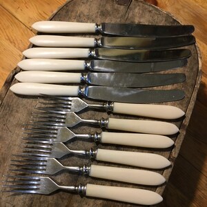 May include: Set of eight silver-plated knives and forks with ivory-colored handles. The knives and forks are arranged in a row on a wooden surface.