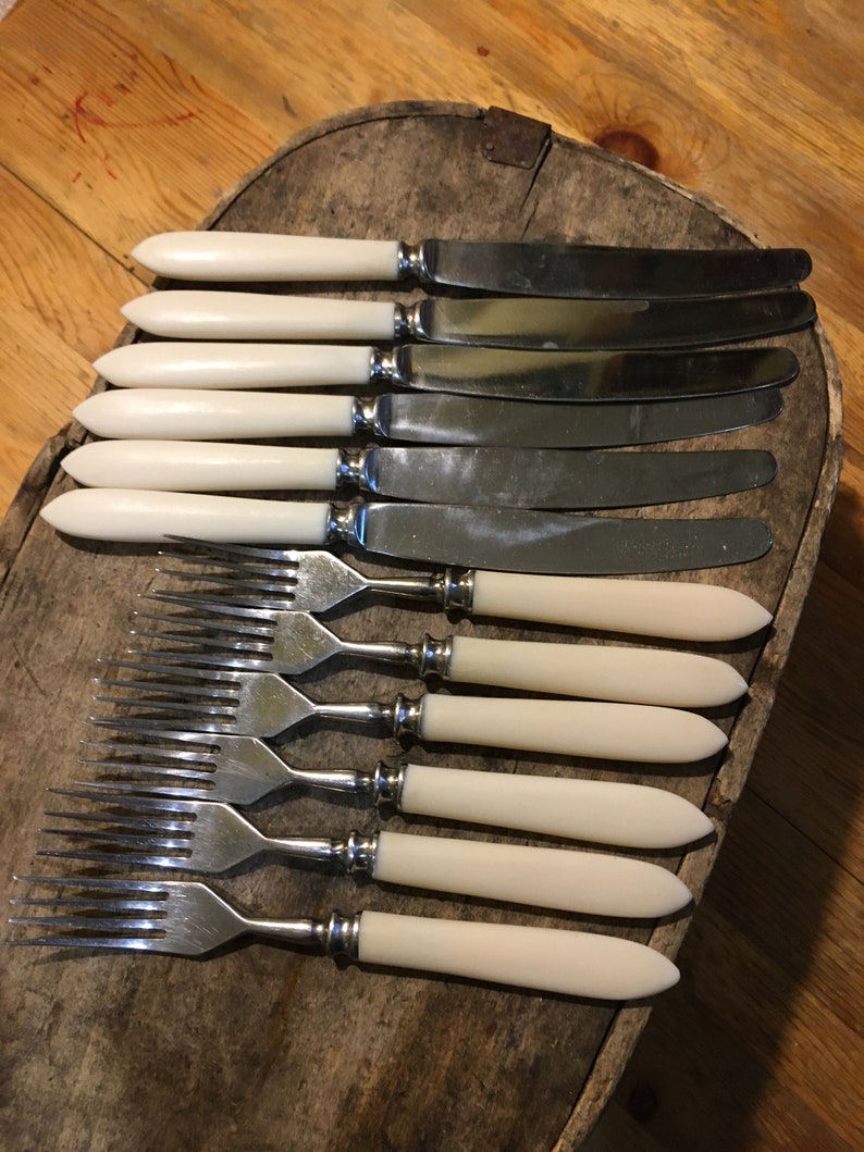 May include: Set of eight silver-plated knives and forks with ivory-colored handles. The knives and forks are arranged in a row on a wooden surface.