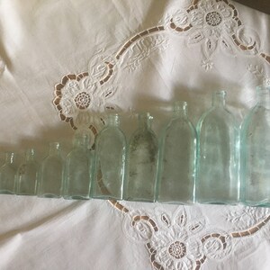 Puede incluir: Una colecci&oacute;n de nueve botellas de vidrio transparente dispuestas en fila de menor a mayor, con una regla blanca colocada verticalmente junto a la botella m&aacute;s grande para mostrar la escala. Las botellas tienen una base rectangular y un cuello corto y estrecho.