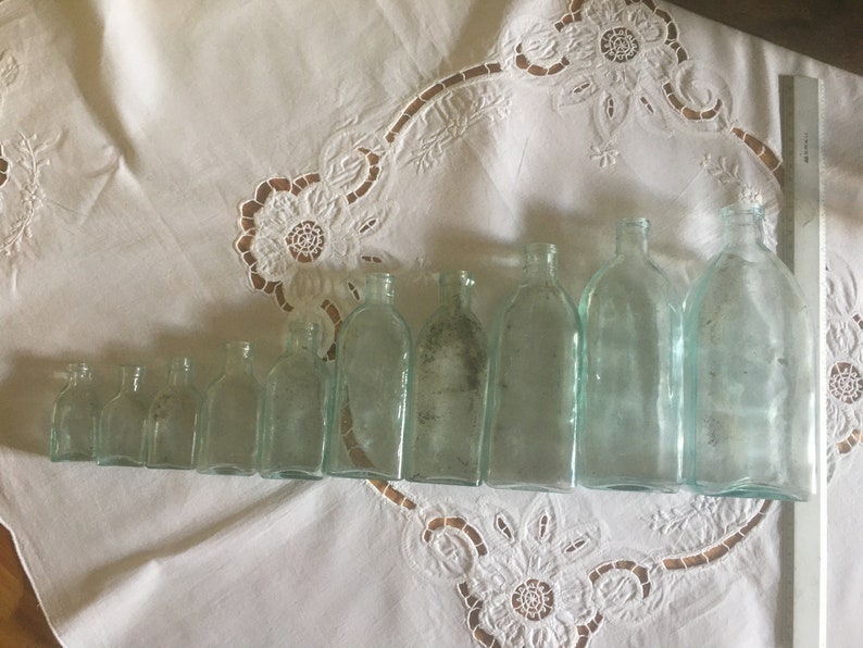 Puede incluir: Una colecci&oacute;n de nueve botellas de vidrio transparente dispuestas en fila de menor a mayor, con una regla blanca colocada verticalmente junto a la botella m&aacute;s grande para mostrar la escala. Las botellas tienen una base rectangular y un cuello corto y estrecho.