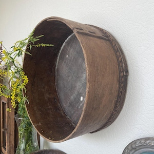 May include: A large, antique wooden sieve with a dark brown finish. The sieve is round with a mesh bottom and is mounted on a white wall. The wooden frame shows signs of age and wear, adding to its rustic charm. The sieve is a decorative piece.