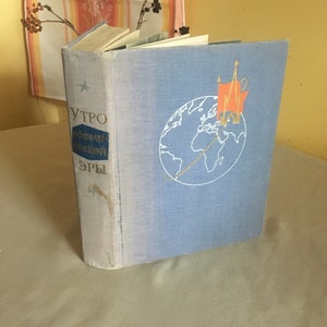 Puede incluir: Un libro de tapa dura azul con el título "УТРО КОСМИЧЕСКОЙ ЭРЫ" en el lomo. La portada presenta un globo terráqueo con una estrella roja y un edificio en la cima.