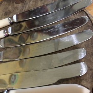 May include: Set of six silver butter knives with white handles. The knives are arranged in a row, with the handles facing the viewer. The knives are shiny and reflect the light.