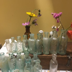 May include: A collection of small, clear glass bottles with a variety of flowers in them. The bottles are arranged in a row on a wooden surface. The flowers are yellow, pink, and purple.