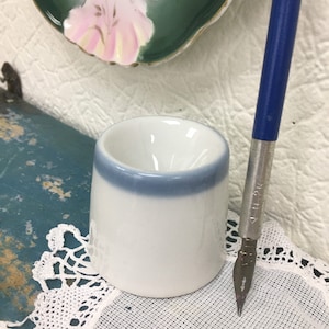 Può includere: Un portapenne in ceramica bianca con una fascia blu intorno alla parte superiore. Il portapenne è su un pizzo bianco all'uncinetto. Una penna blu e argentata è accanto al portapenne.