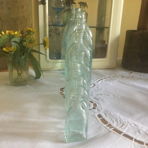 Puede incluir: Una colecci&oacute;n de botellas de vidrio transparente con un ligero tono aguamarina, dispuestas en una fila. Cada botella presenta un cuerpo redondeado, un cuello corto y una peque&ntilde;a abertura circular en la parte superior. El vidrio tiene una apariencia lisa y ligeramente transl&uacute;cida.