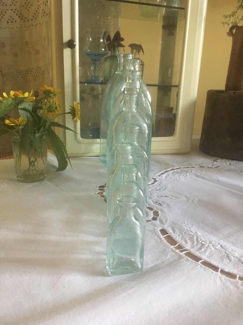 Puede incluir: Una colecci&oacute;n de botellas de vidrio transparente con un ligero tono aguamarina, dispuestas en una fila. Cada botella presenta un cuerpo redondeado, un cuello corto y una peque&ntilde;a abertura circular en la parte superior. El vidrio tiene una apariencia lisa y ligeramente transl&uacute;cida.