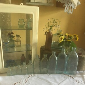 Puede incluir: Un armario de pared blanco con puerta de cristal que contiene varios frascos de vidrio peque&ntilde;os, junto a una colecci&oacute;n de botellas de vidrio azul claro transparente de tama&ntilde;os graduados en fila, y una jarra de cer&aacute;mica marr&oacute;n con flores amarillas.