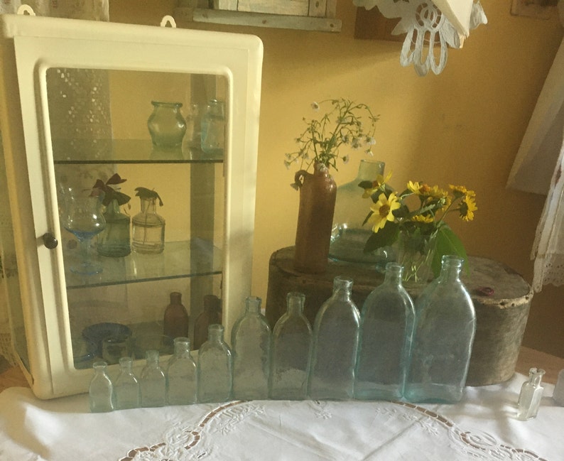 Puede incluir: Un armario de pared blanco con puerta de cristal que contiene varios frascos de vidrio peque&ntilde;os, junto a una colecci&oacute;n de botellas de vidrio azul claro transparente de tama&ntilde;os graduados en fila, y una jarra de cer&aacute;mica marr&oacute;n con flores amarillas.