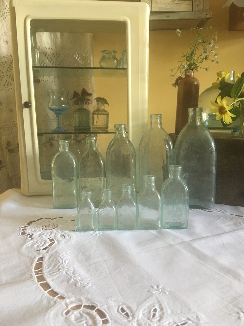 Puede incluir: Una colecci&oacute;n de diez botellas de vidrio transparente y de tono azul claro de diversos tama&ntilde;os y formas, dispuestas en dos filas. Las botellas presentan cuellos estrechos y cuerpos redondeados, mostrando algunas texturas superficiales sutiles.