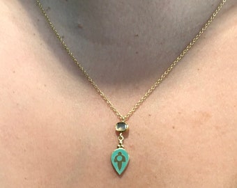 Op de afbeelding: Een gouden ketting met een hanger in de vorm van een tealblauwe traan met een gouden figuur erin en een kleine bruine edelsteen erboven.