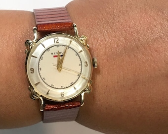 May include: A gold-tone wristwatch with a white face and brown leather band. The watch face has black Roman numeral hour markers and a red, white, and blue logo that says "BENRUS".